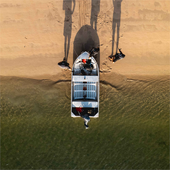 Aerial top-down view of a white Quintrex Explorer being pushed into the water from a sandy beach by two people.
