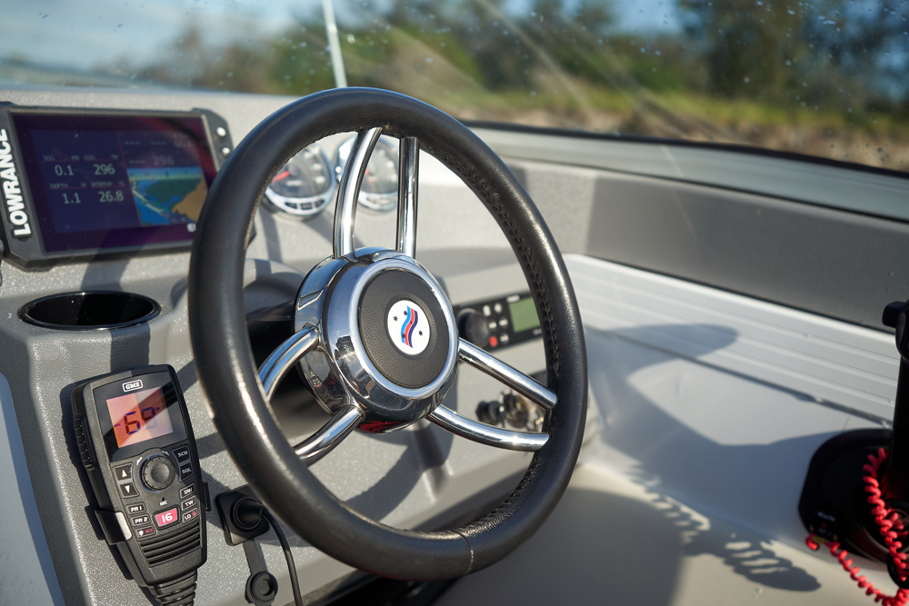 Close-up of a Quintrex boat helm featuring a three-spoke steering wheel, digital navigation display, and a VHF radio.