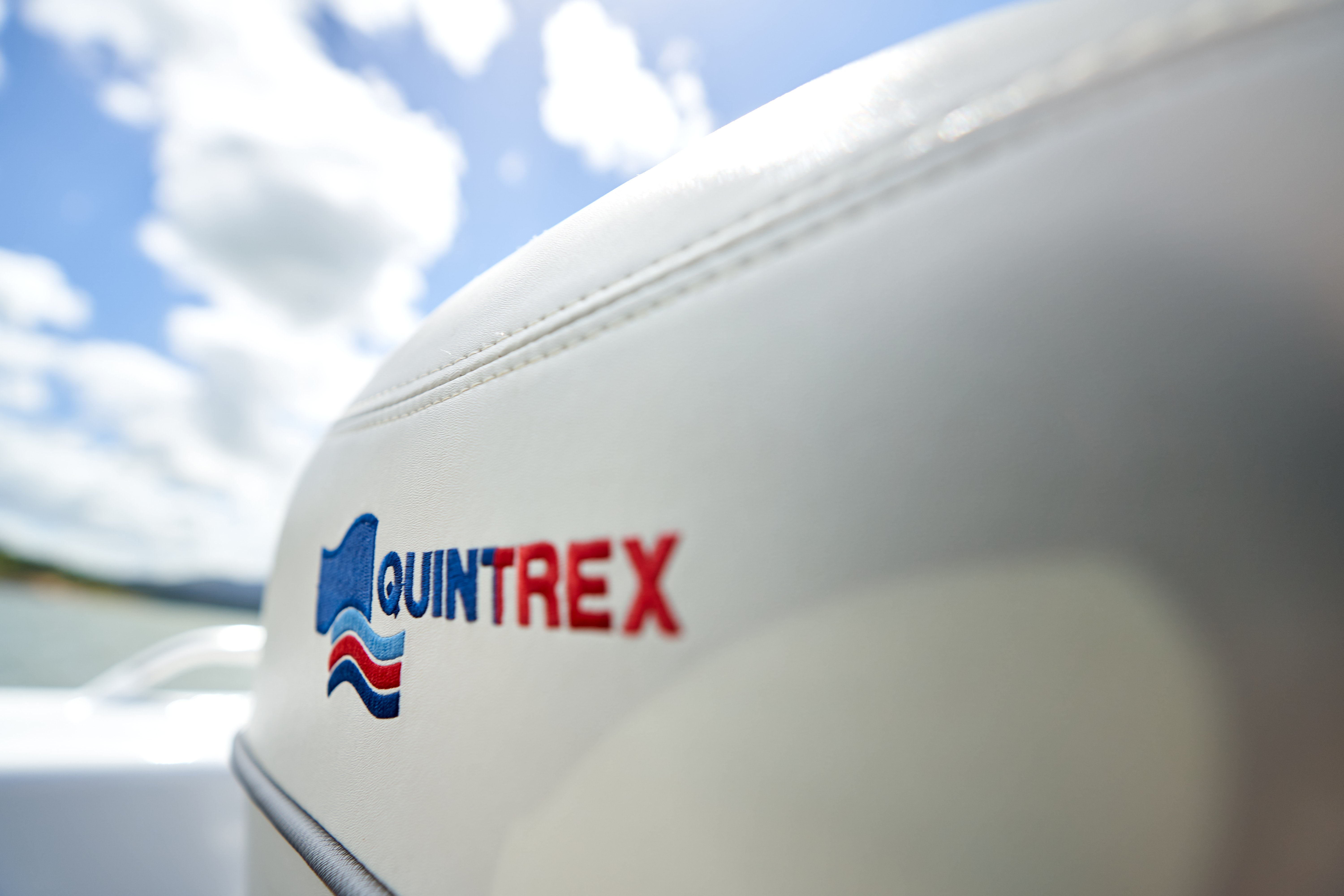 Close-up of a white Quintrex boat seat featuring the embroidered logo against a blurred sky and water background.
