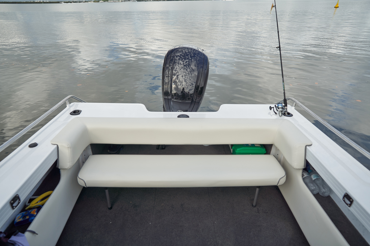 Rear view of a Quintrex boat's white cushioned bench seat, outboard motor, and a fishing rod overlooking calm water.