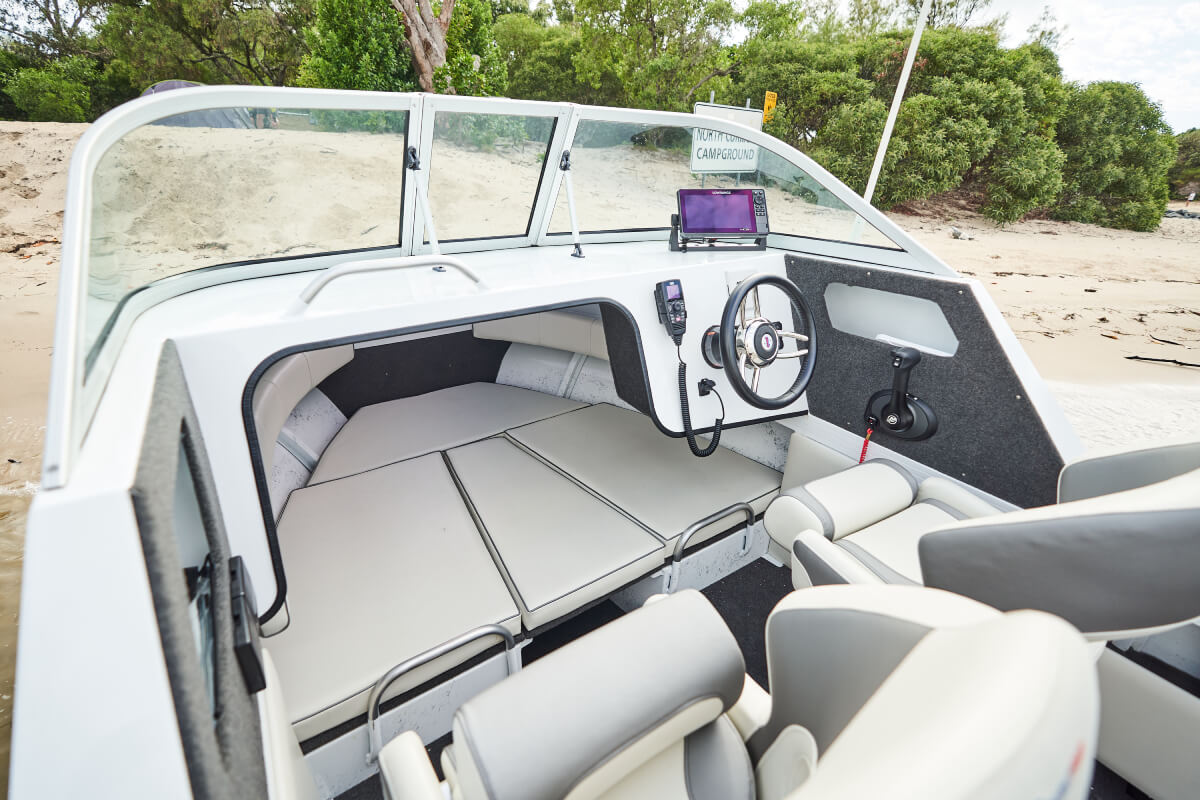 The interior of a white Quintrex cabin boat featuring cushioned seating, a steering wheel, and a dashboard near a sandy beach.