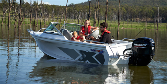 A family in life jackets navigates a Quintrex Fishabout through a lake with bare trees and a mountain backdrop.